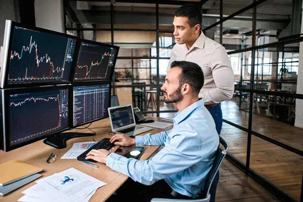 Two traders studying candlestick charts on multiple monitors in an office