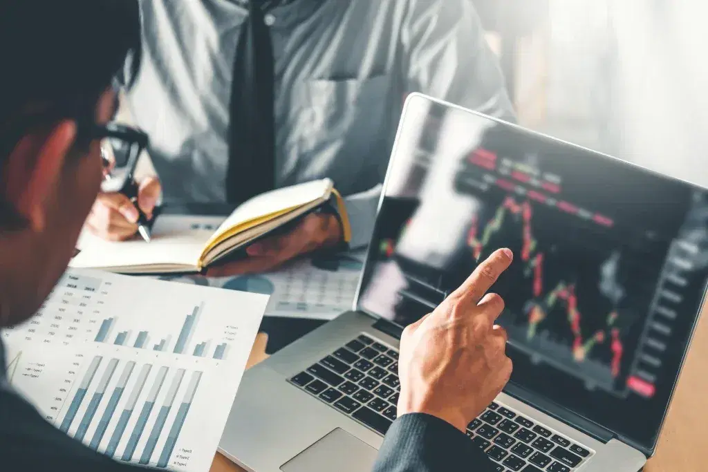 Trader pointing at a candlestick chart on a laptop beside printed bar charts